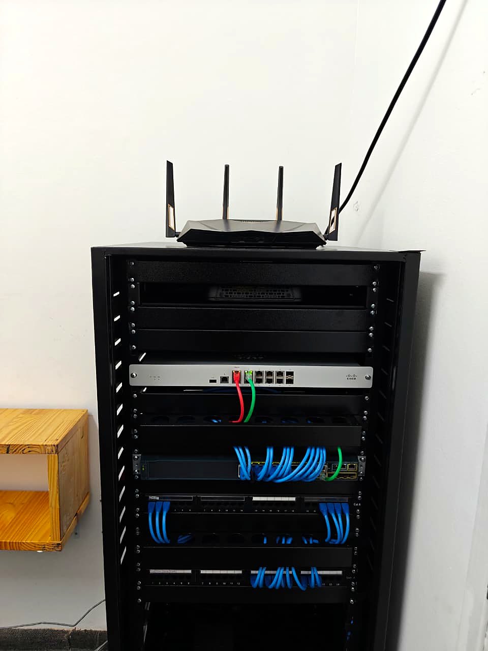 Vista frontal do rack finalizado com roteador no topo, firewall, switch e patch panels conectados por patch cords azuis