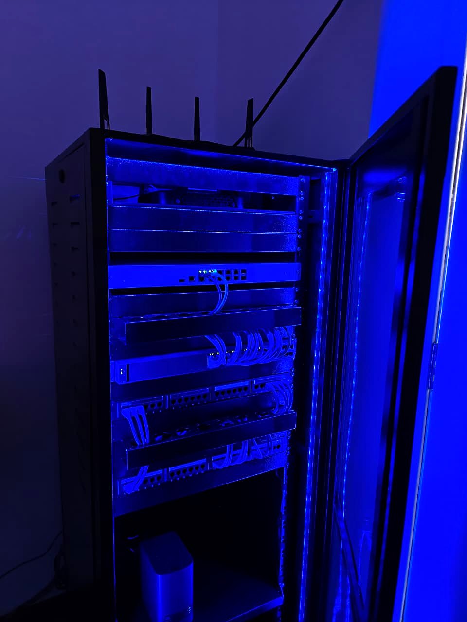 Rack com a porta de vidro aberta e iluminação LED azul interna, com firewall, switch e patch panels em cascata