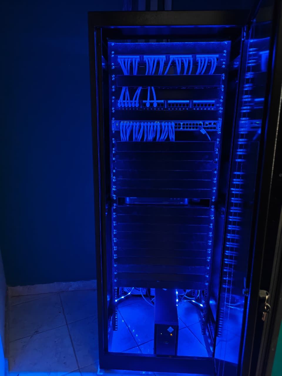 Vista frontal completa do rack com a porta entreaberta, equipamentos organizados, painéis cegos e nobreak na base
